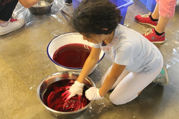 Mädchen hockt auf dem Boden vor zwei Gefäßen und färbt ein Kleidungsstück rot ein.