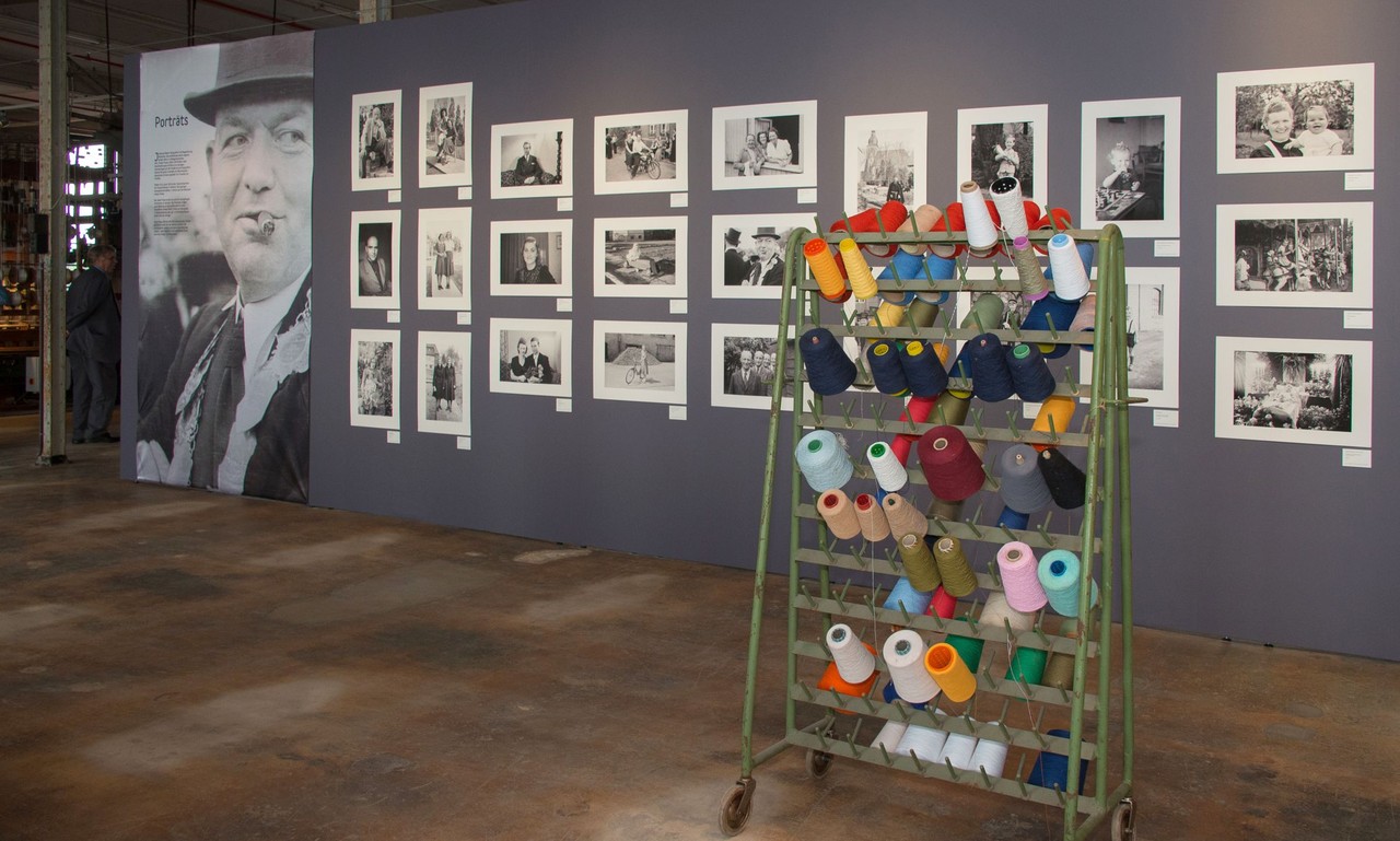 Blick in die Ausstellung "Lust auf Leben" mit Schwarz-Weiß-Fotografien an der Wand. Im Vordergrund ein Ständer mit farbigen Garnspulen.