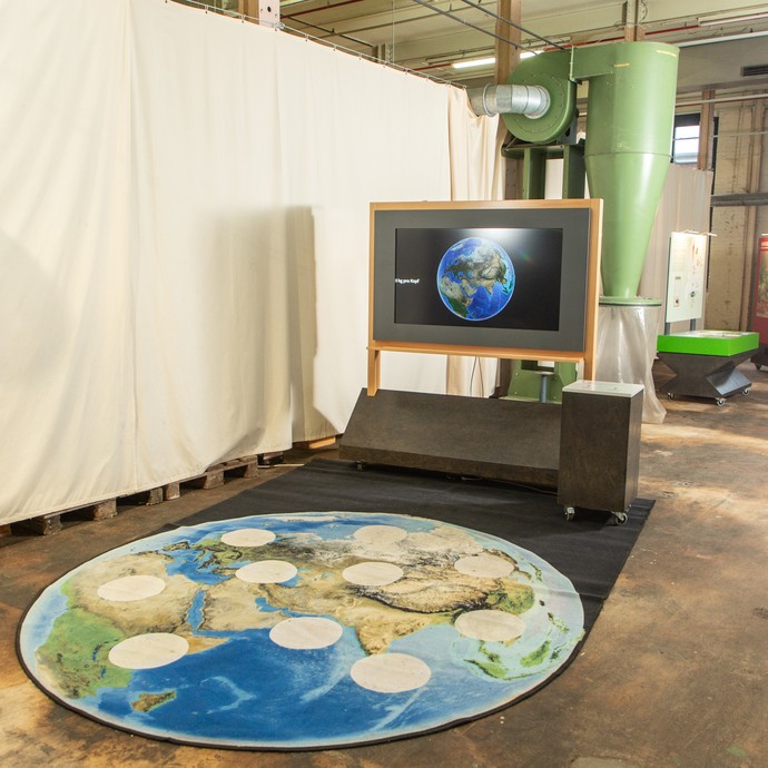 Blick in die Ausstellung: Abbildung einer Weltkugel auf dem Boden vor einer Medienstation (vergrößerte Bildansicht wird geöffnet)