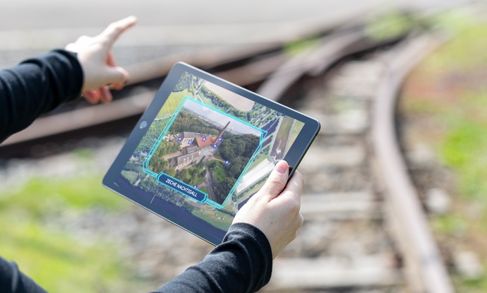 Blick auf ein Tablet in den Händen einer Besucherin. Im Hintergrund sind Gleise der Zeche Nachtigall zu sehen. Blick auf ein Tablet in den Händen einer Besucherin. Im Hintergrund sind Gleise der Zeche Nachtigall zu sehen.