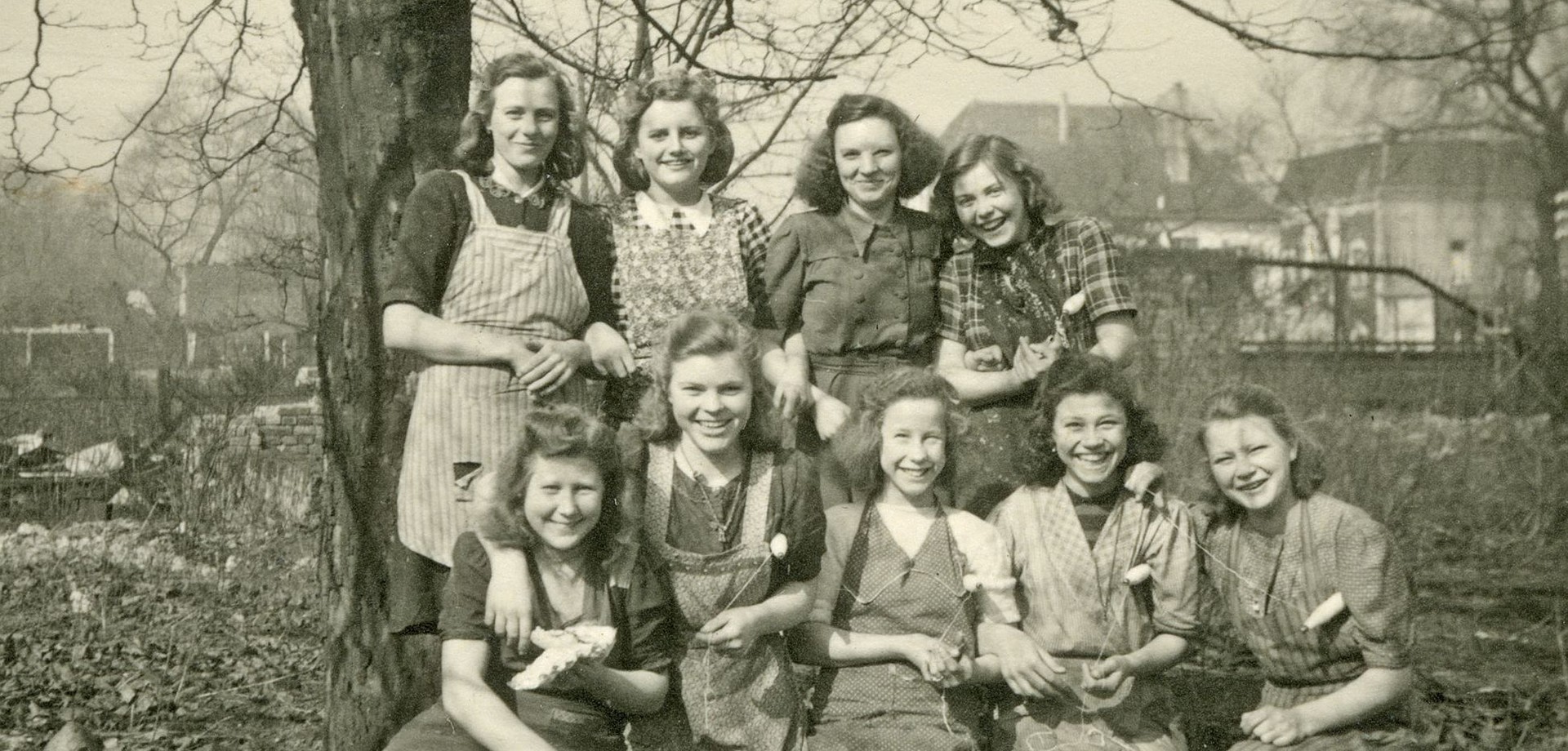 Schwarz-Weiß Aufnahme mit 9 Mitarbeiterinnen der Spinnerei Herding. Im Hintergrund sind Bäume und Gebäude zu sehen.