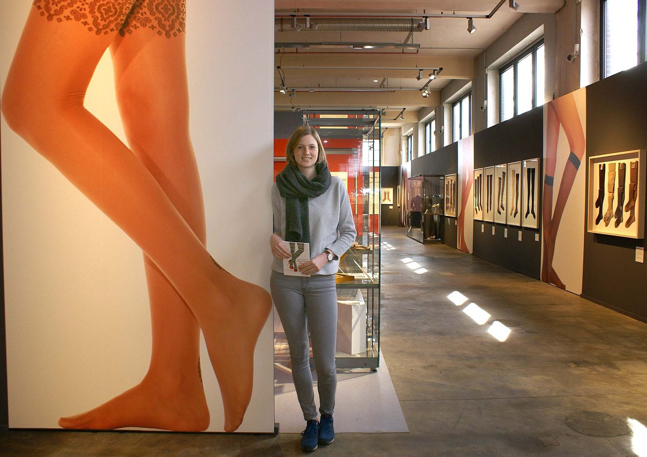 Kuratorin Christa Frins steht neben einm großen Foto, das Frauenbeine in Nylonstrümpfen zeigt. Dahinter öffnet sich der Blick in die Ausstellung.